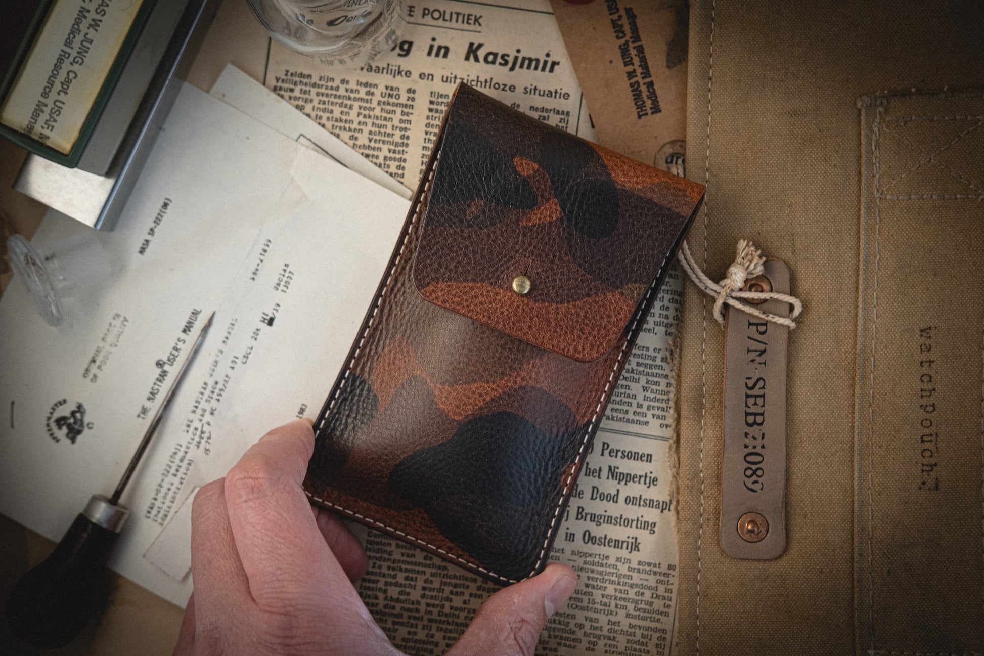 A hand holds a pouch from the Camo veg-tan collection. Below are various papers and a foreign language newspaper clipping. To the right, a canvas watch pouch with a "WATCHPOUCH" leather tag rests, all intricately handmade in Antwerp.