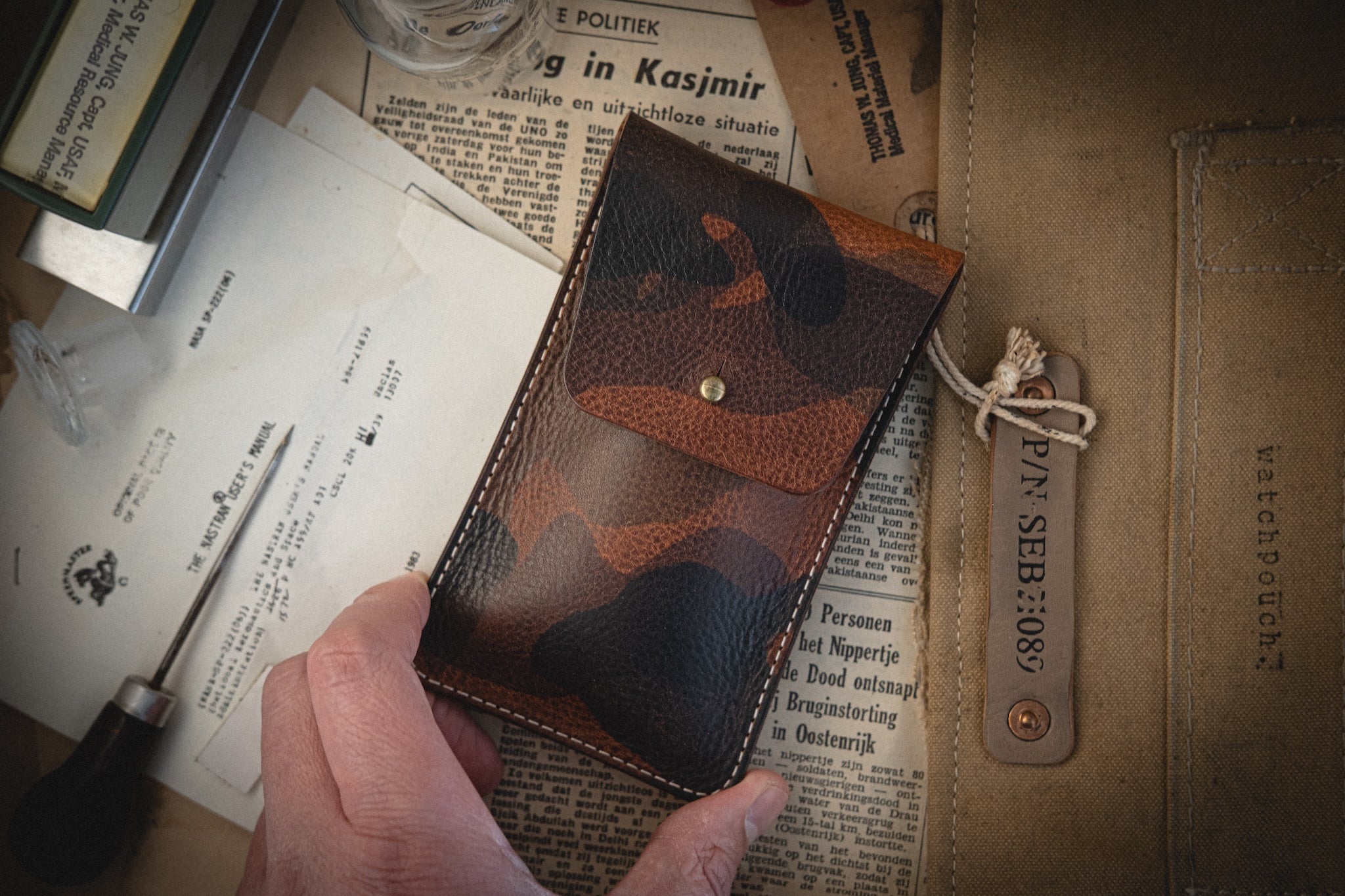 A hand holds a pouch from the Camo veg-tan collection. Below are various papers and a foreign language newspaper clipping. To the right, a canvas watch pouch with a "WATCHPOUCH" leather tag rests, all intricately handmade in Antwerp.