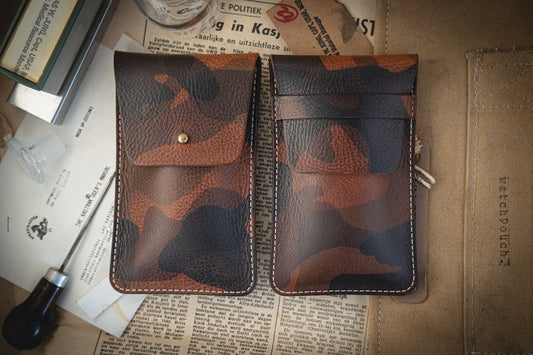 Two pouches from the Camo veg-tan collection, handmade in Antwerp, sit on a vintage table with scattered papers. One has a snap button; the other features a strap with an attached key, enhancing the rustic charm of the setting.