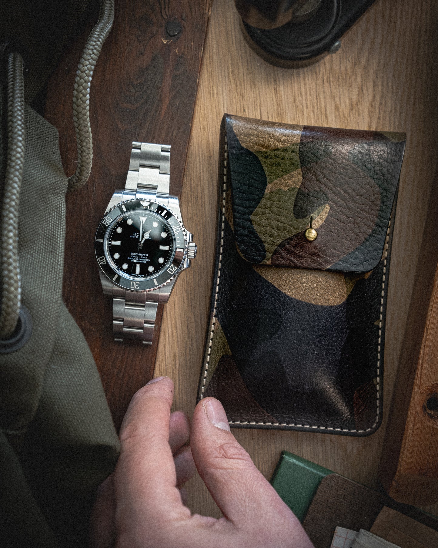 A hand holds a rectangular pouch from the Veg-tan CAMO COLLECTION on a wooden surface. Next to it lies a stainless steel wristwatch with a black dial and bezel. The image also features parts of a green backpack and other indistinct objects, including what seems to be a microfiber cleaning cloth peeking out.