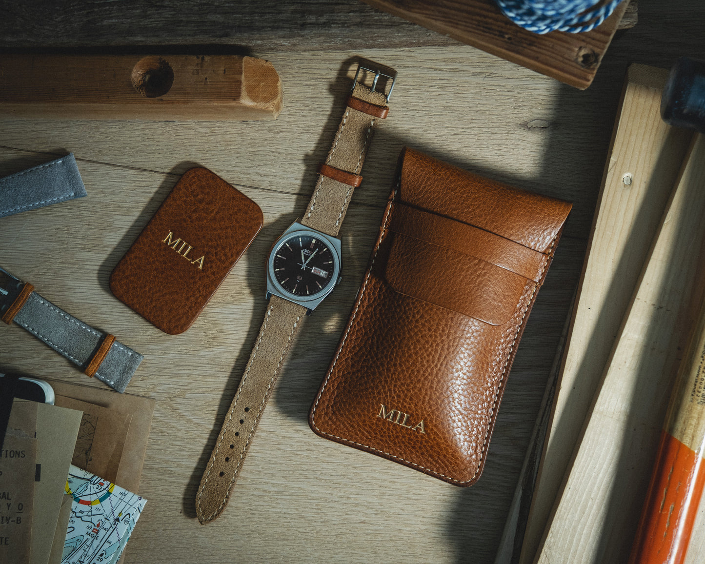 A flat lay of the Pebbled veg-tan collection "Earth tones" featuring a brown leather watch with a silver face, placed alongside a matching leather pouch and cardholder embossed with "MILA." The items are artistically arranged on a wooden surface surrounded by tools and accessories, emanating warm Earth tones and a personalized touch.