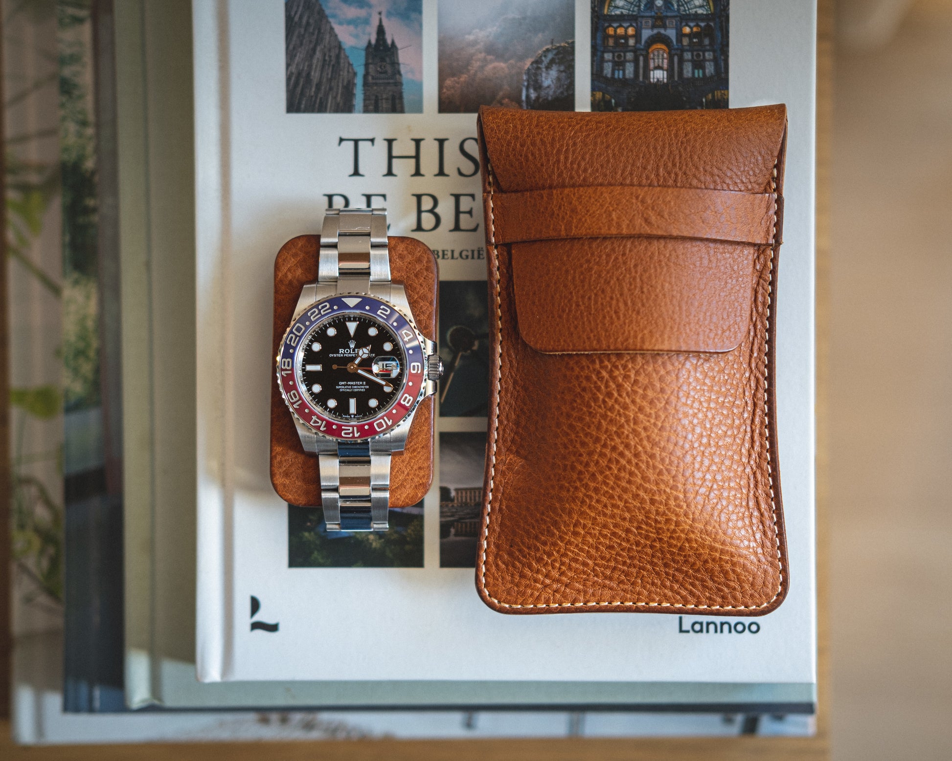 A stylish wristwatch with a red and blue bezel, black dial, and silver strap rests on a few books. Next to the watch is a brown leather pouch from the Pebbled veg-tan collection "Earth tones." The books, adorned with images of architecture on their covers and titles partially visible, add an elegant touch of Earth tones to the scene.