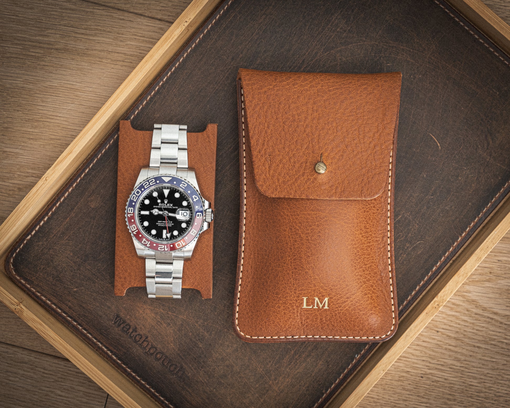 A stainless steel wristwatch with a black dial and multicolored bezel is displayed on a leather watch stand. Next to it, there is a brown pebbled veg-tan leather watch pouch from the "Earth tones" collection, monogrammed with "LM" for personalization. Both items are arranged on a wooden tray featuring a dark leather base that complements the Earth tones.