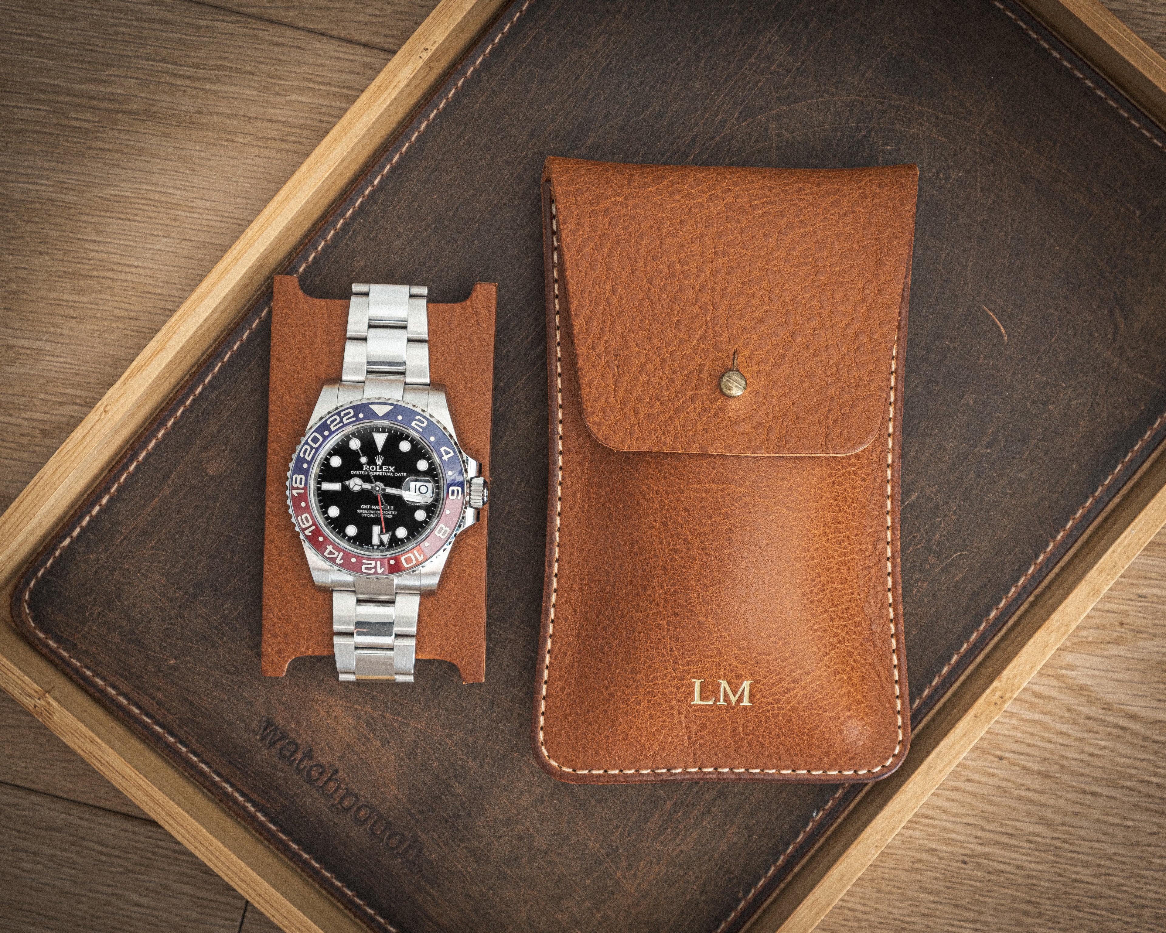 A stainless steel wristwatch with a black dial and multicolored bezel is displayed on a leather watch stand. Next to it, there is a brown pebbled veg-tan leather watch pouch from the "Earth tones" collection, monogrammed with "LM" for personalization. Both items are arranged on a wooden tray featuring a dark leather base that complements the Earth tones.