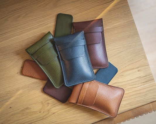 An assortment of pebbled veg-tan leather pouches from the "Earth tones" collection is arranged on a wooden surface. The pouches, available in green, navy blue, brown, and tan, each feature a card slot on the front and a flap at the top. The lighting beautifully highlights the texture of the leather.