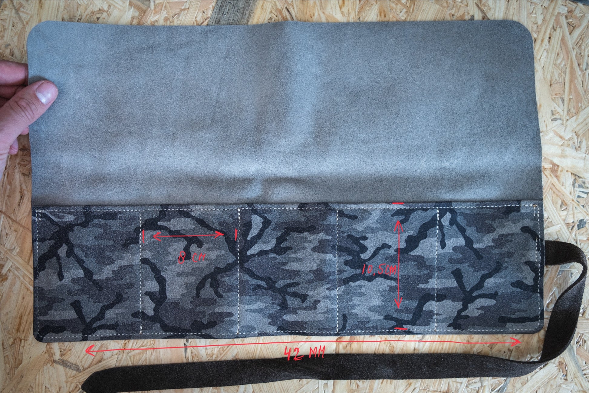 A hand opens a camouflage-patterned "SAMPLE SALE watchroll Camo grey 5 pocket" on a wooden surface. The suede case includes several pockets with red measurement markings, with one measuring 8 cm wide and another 10.5 cm in height, and the total width of the case is 42 cm.
