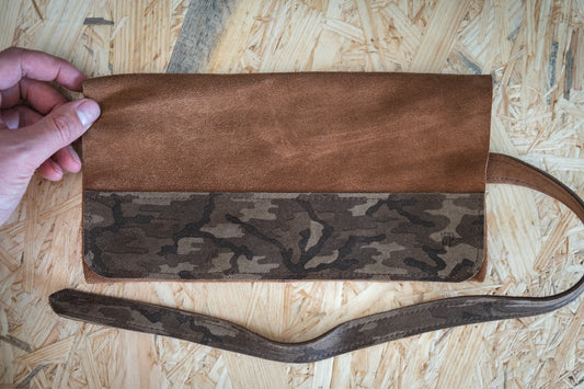 A hand holding a brown leather clutch with a camouflage-patterned strip along the bottom rests on a wooden surface, accompanied by a sleek SAMPLE SALE watchroll suede brown camo 4 pocket.