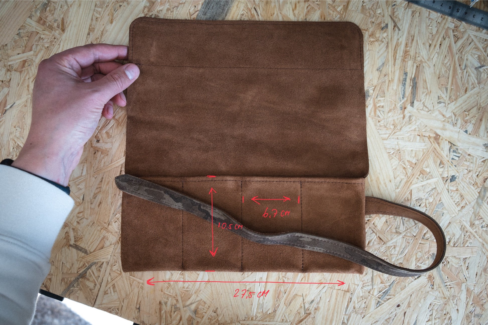 On a wooden surface, someone holds open the SAMPLE SALE watchroll in brown camo suede. Red measurements of 6.9 cm, 10.5 cm, and 23.5 cm are drawn on it, and it features a strap attached to the side.