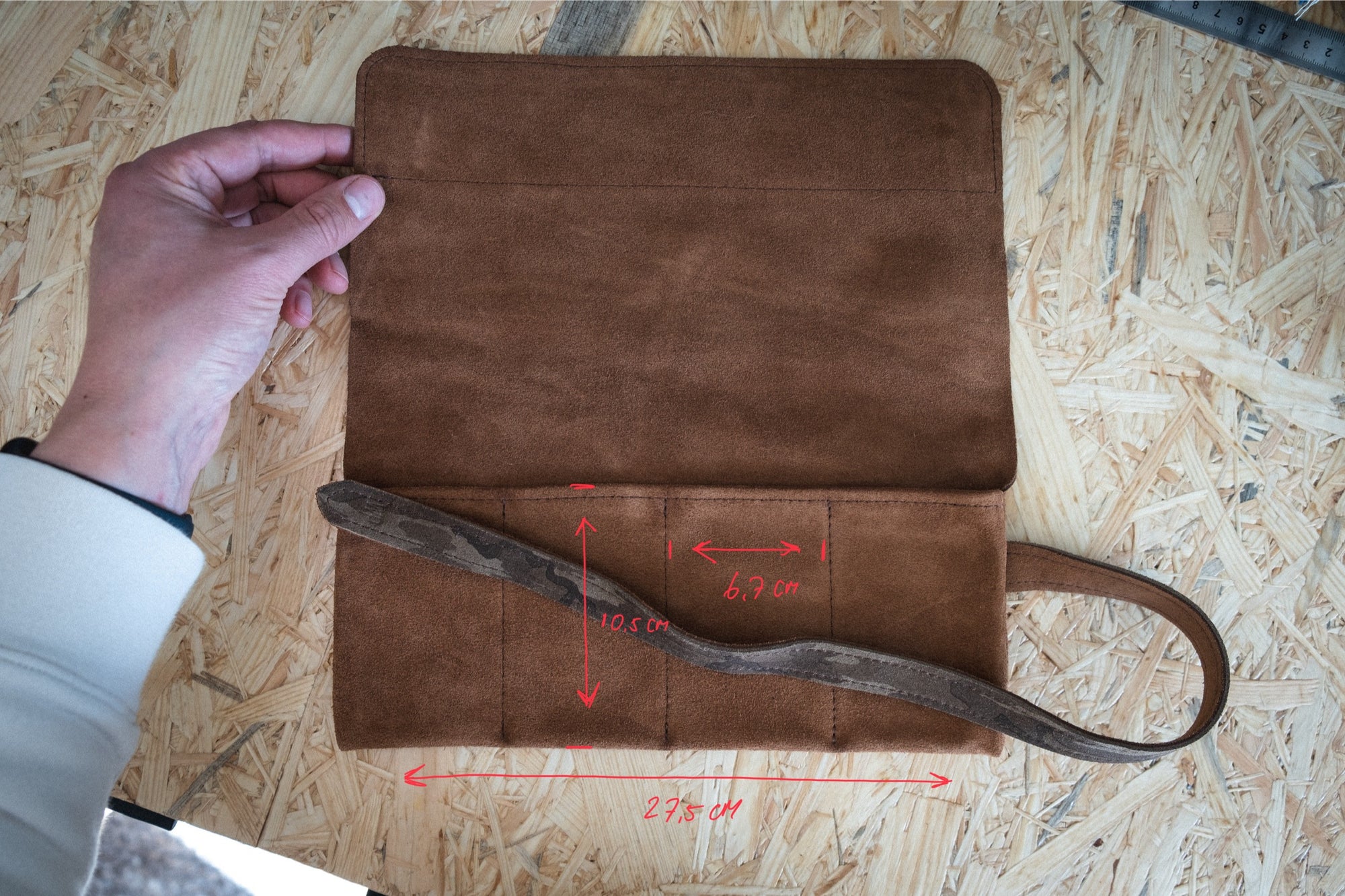 On a wooden surface, someone holds open the SAMPLE SALE watchroll in brown camo suede. Red measurements of 6.9 cm, 10.5 cm, and 23.5 cm are drawn on it, and it features a strap attached to the side.