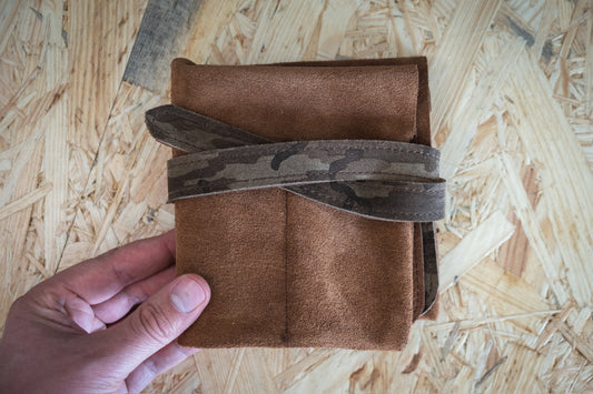 A hand holds the SAMPLE SALE watchroll suede brown camo with 4 pockets, featuring a camouflage strap artfully tied around it. The background showcases a light-colored, textured wooden surface.