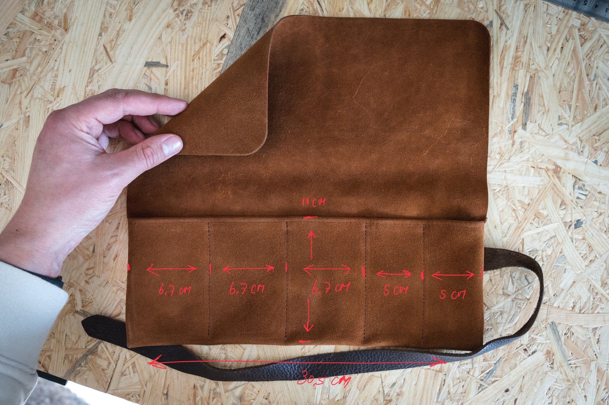 A person displays a SAMPLE SALE watchroll suede brown camo 5 pocket, featuring five suede-lined compartments each labeled with measurements. The watchroll measures 30.5 cm in width and 11 cm in height and is placed on a wooden surface.