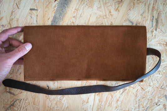 A hand holding a SAMPLE SALE watchroll in brown suede camo with five pockets and a dark leather strap rests elegantly on a textured wooden surface.