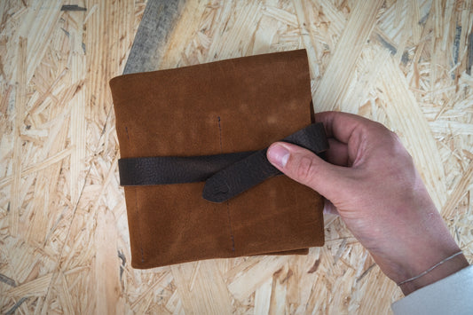 A hand holds a luxuriously textured SAMPLE SALE watchroll in brown suede camo with five pockets on a light wooden surface, its rich appearance reminiscent of finely crafted leather.
