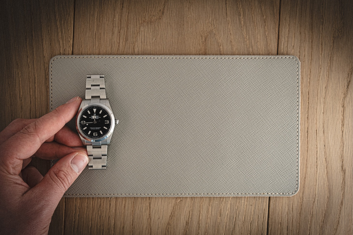A hand holds a silver wristwatch with a black face and white numbers, elegantly positioned on the SAMPLE SALE Valet 13 * 23 cm made of grey saffiano leather atop a textured beige mat on the wooden surface.
