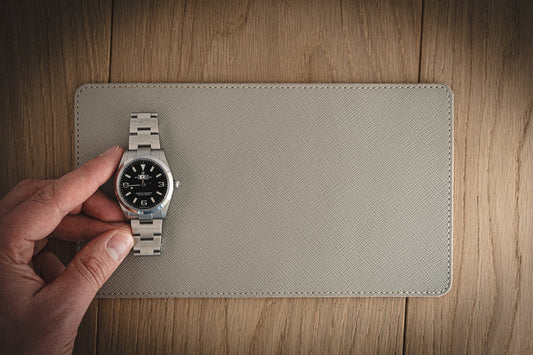 A hand holds a silver wristwatch with a black face and white numbers, elegantly positioned on the SAMPLE SALE Valet 13 * 23 cm made of grey saffiano leather atop a textured beige mat on the wooden surface.