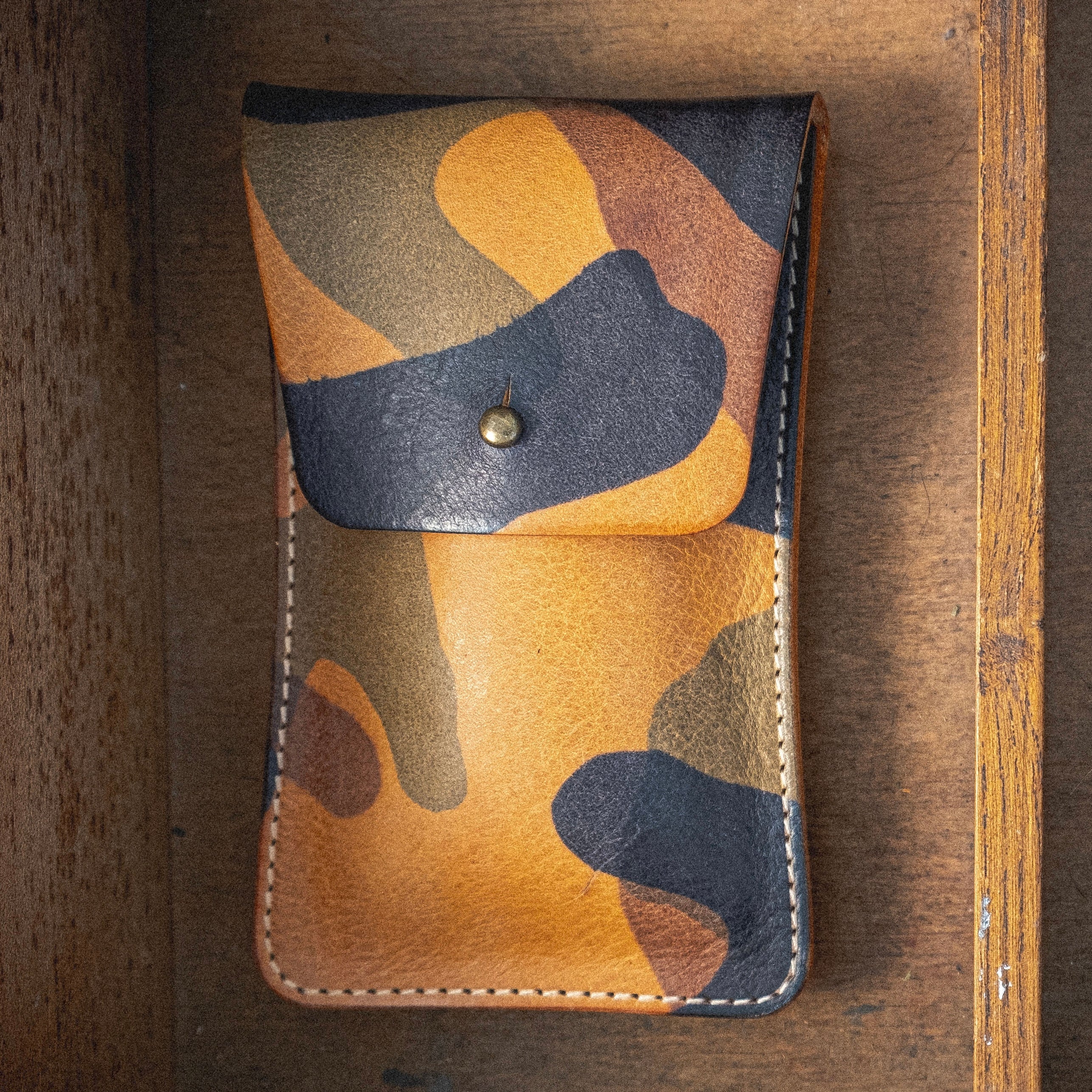 The Veg-tan CAMO COLLECTION pouch, featuring a camouflage pattern and equipped with a snap button closure, is displayed against a wooden surface. This handmade watch pouch, blending green, brown, and tan hues, exudes a rugged and natural aesthetic.