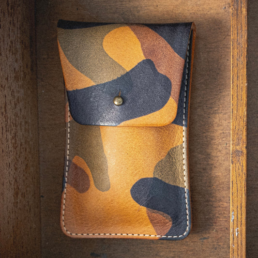 The Veg-tan CAMO COLLECTION pouch, featuring a camouflage pattern and equipped with a snap button closure, is displayed against a wooden surface. This handmade watch pouch, blending green, brown, and tan hues, exudes a rugged and natural aesthetic.