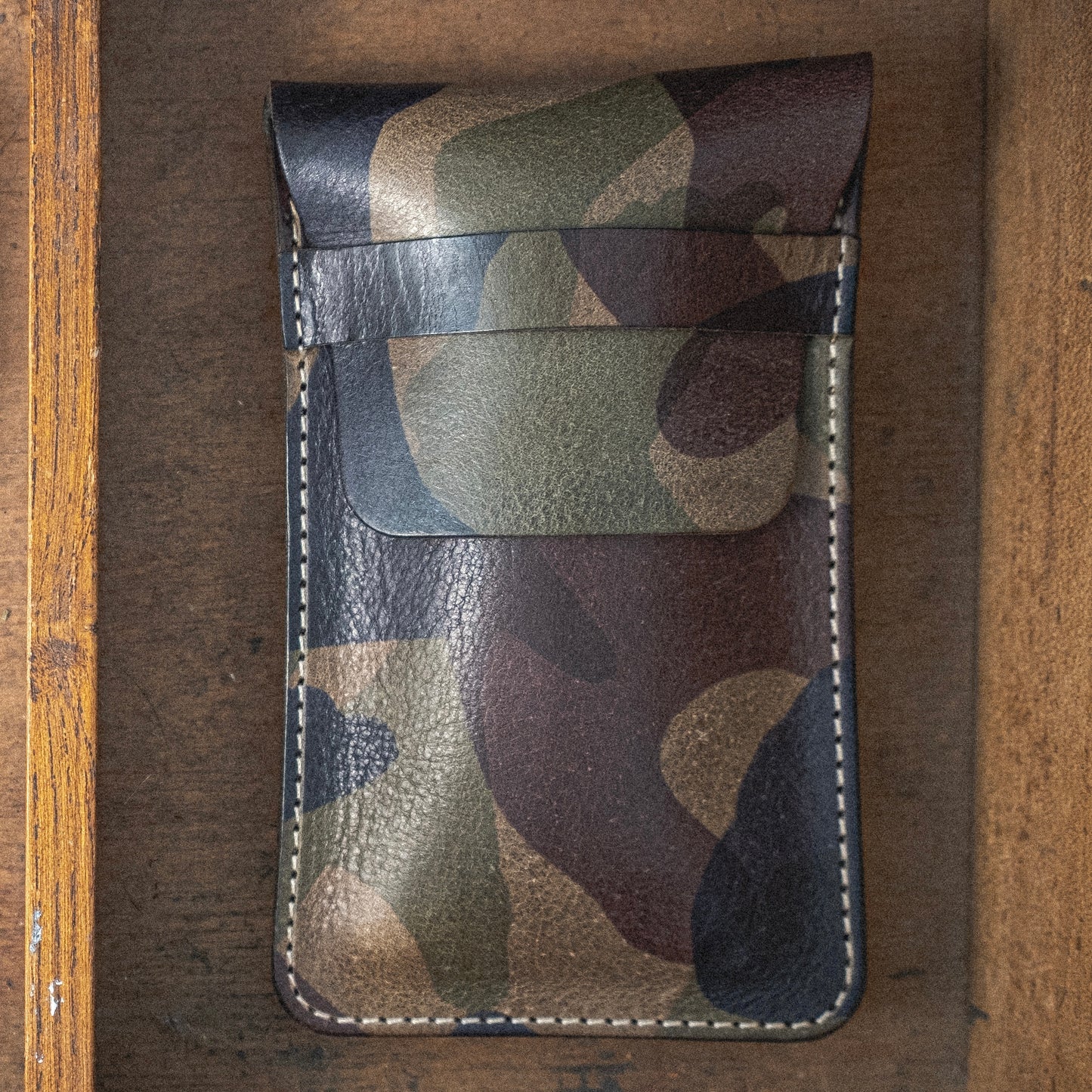 The Veg-tan CAMO COLLECTION leather wallet, featuring a striking camouflage pattern, rests on a wooden surface. This meticulously crafted wallet has stitched edges and a top flap for closure. The background accentuates the wood's distinctive grain and texture.