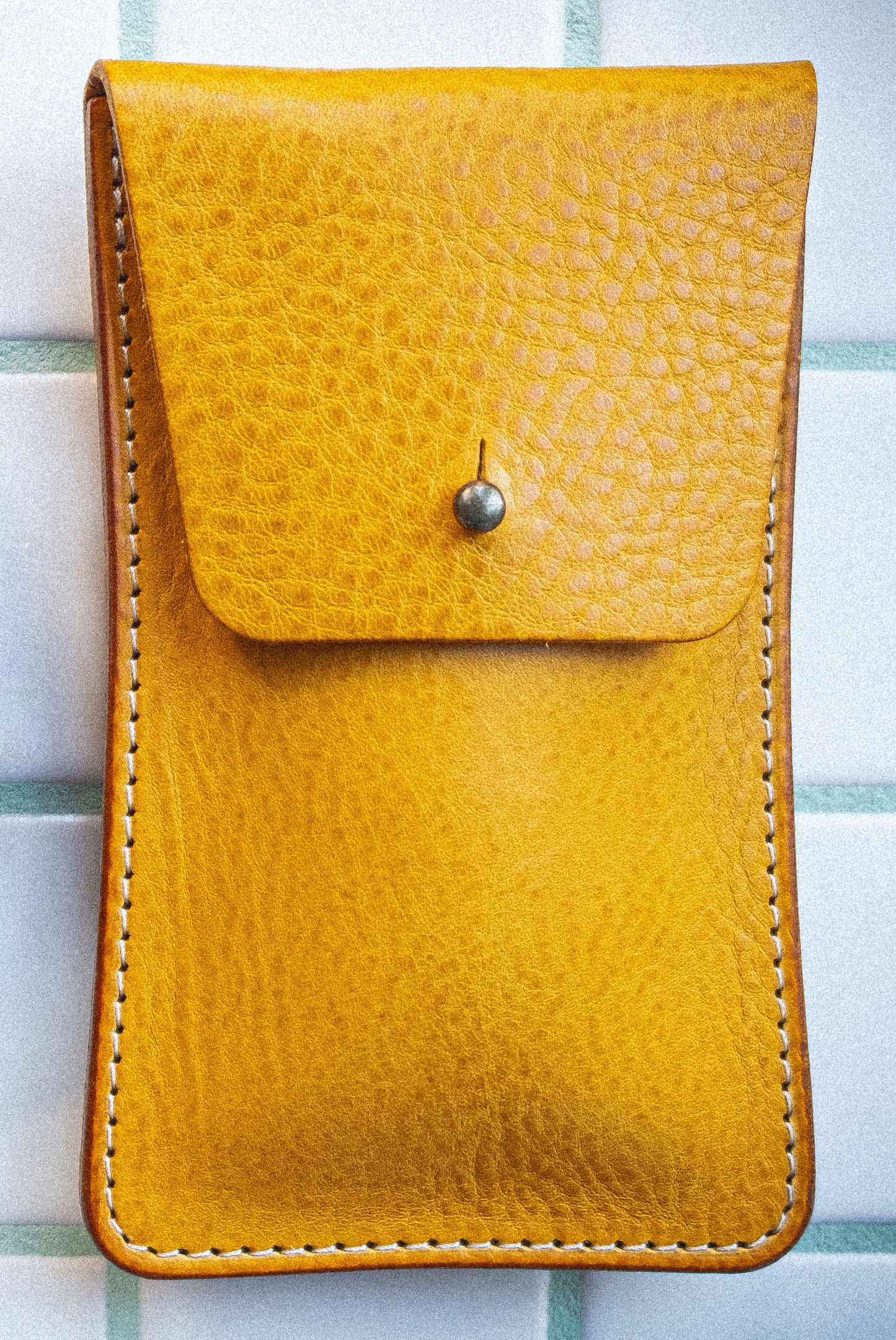 A close-up of the "Vibrant" pouch from the Pebbled Veg-Tan Collection, handmade in Antwerp with mustard yellow leather, features a flap secured by a small metal button. The pouch has visible stitching along the edges and is set against a light-colored, tiled background.