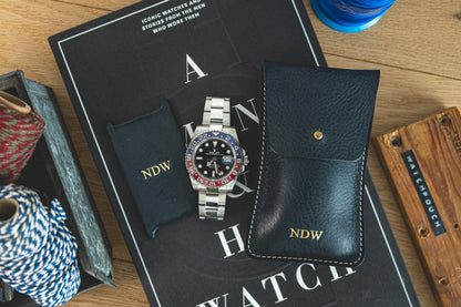 A silver wristwatch with a red and blue bezel is displayed on a book titled "A Man & His Watch." The watch comes with a black pouch and a black leather case, both inscribed with “NDW.” Partially visible on the sides are colored cords and a wooden box from the Pebbled veg-tan collection "Earth tones.