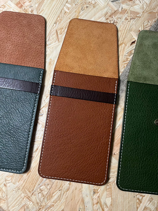 Three SAMPLE SALE veg tan wallets, in cognac and dark brown straps, are displayed open on a wooden surface, showcasing their inner compartments and distinct textures. These sample-made wallets offer a touch of personalization, perfect for those seeking a unique leather accessory.