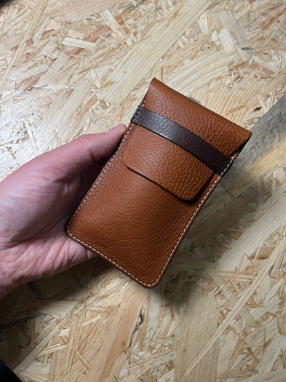 A hand holds the SAMPLE SALE veg tan leather wallet in cognac, featuring a dark brown strap and visible stitching. This personalized leather case protector is displayed against a textured, light-colored wooden surface, emphasizing its craftsmanship and style.