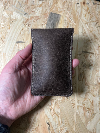 A hand holds the SAMPLE SALE coffee brown premium pouch with its brown strap against a light wooden background. The sleek, rectangular design features visible stitching along the edges, highlighting its craftsmanship.