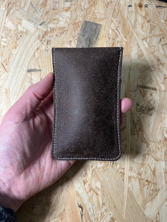 A hand holds the SAMPLE SALE coffee brown premium pouch with its brown strap against a light wooden background. The sleek, rectangular design features visible stitching along the edges, highlighting its craftsmanship.
