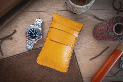 A Pebbled Veg-Tan Collection "Vibrant" wristwatch with a black and red bezel is placed on a wooden surface next to a handmade yellow leather pouch from Antwerp. Surrounding the items are various objects, including leather scraps, a watch case protector, a spool of thread, and a ceramic cup.