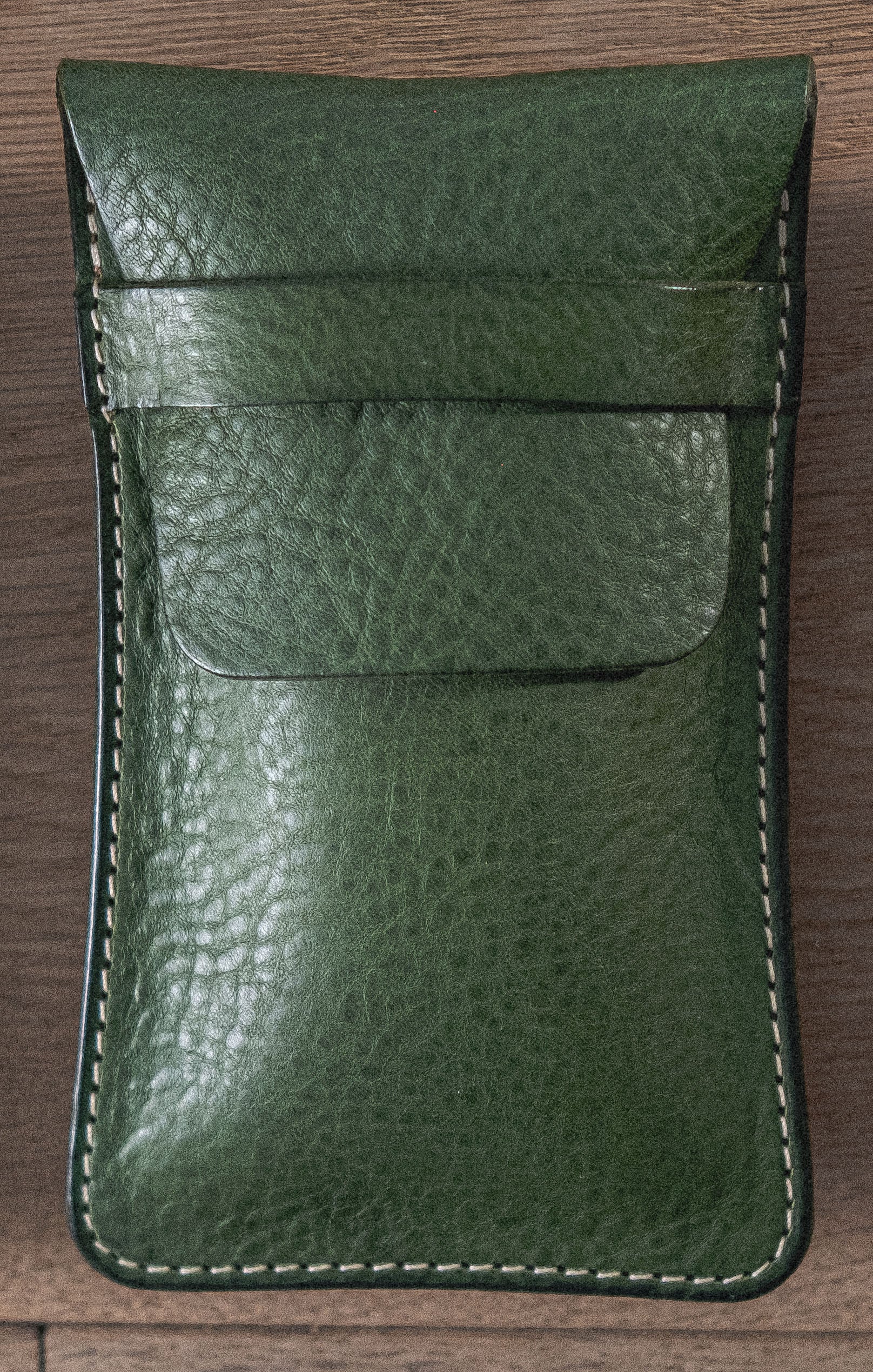 A close-up image of the Pebbled Veg-Tan Collection "Vibrant" green leather pouch with white stitching, handmade in Antwerp. The pouch, crafted from pebbled veg-tan leather, features a flap closure and rests on a wooden surface. The texture of the leather and the details of the stitching are clearly visible.