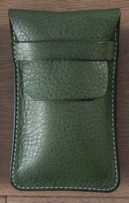 A close-up image of the Pebbled Veg-Tan Collection "Vibrant" green leather pouch with white stitching, handmade in Antwerp. The pouch, crafted from pebbled veg-tan leather, features a flap closure and rests on a wooden surface. The texture of the leather and the details of the stitching are clearly visible.