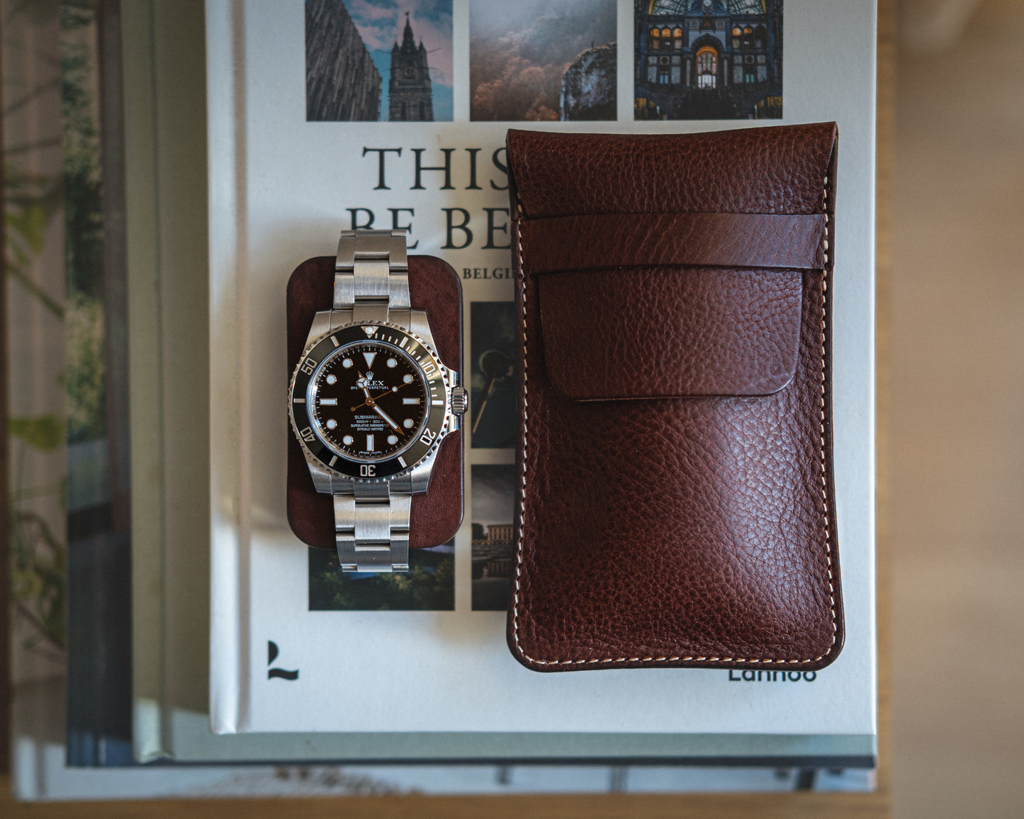 A stainless steel wristwatch with a black dial and bezel sits on top of a stack of books. Next to it is a brown pouch from the Pebbled veg-tan collection "Earth tones." The top book's title reads, "This Is Belgium." The background features other slightly blurred books in rich Earth tones.
