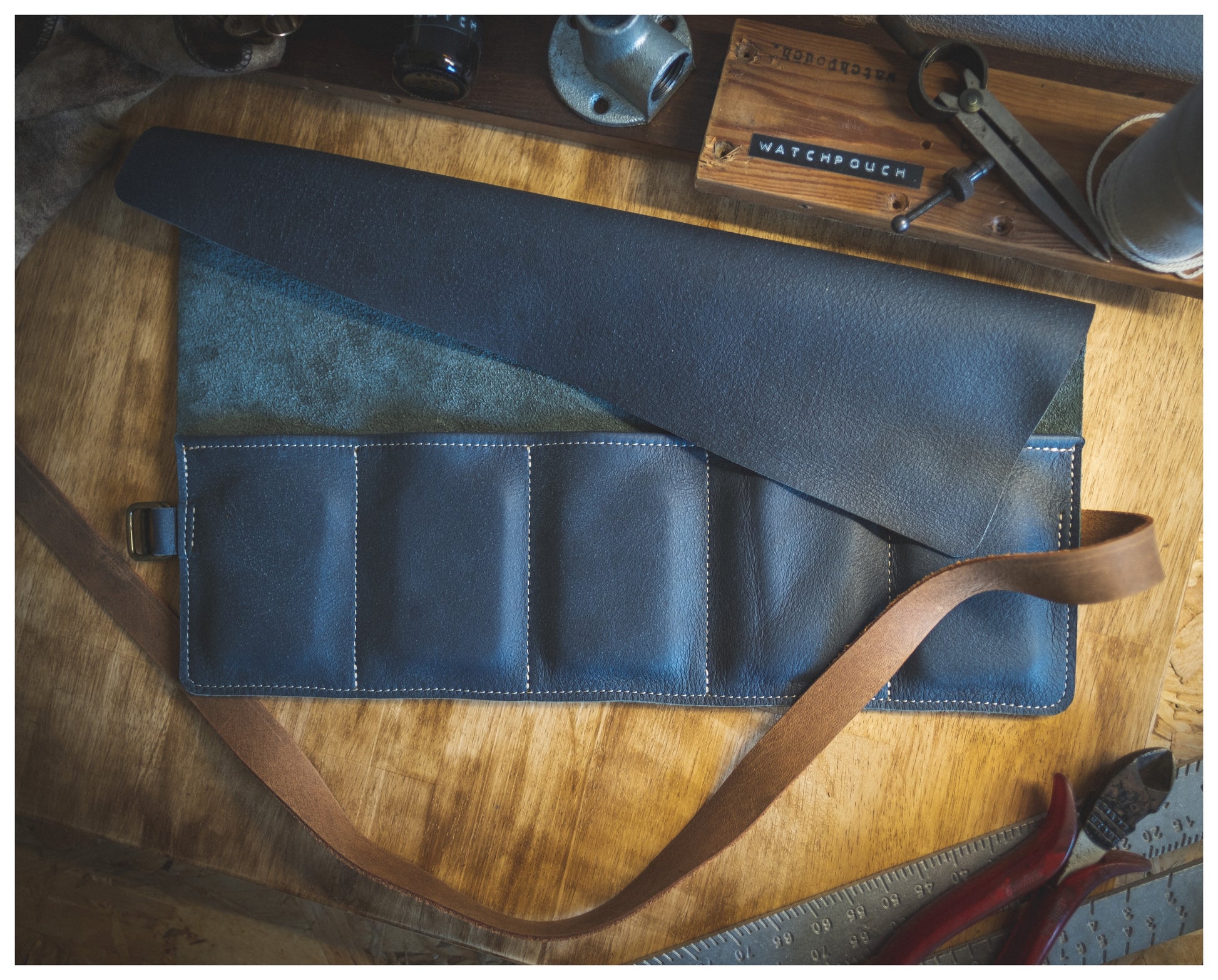 A partially folded watchroll from the Watchroll collection, available in various colors, is laid open on a wooden surface. A personalized brown strap is attached, and various tools and items, such as pliers and a ruler, are scattered around the workspace.