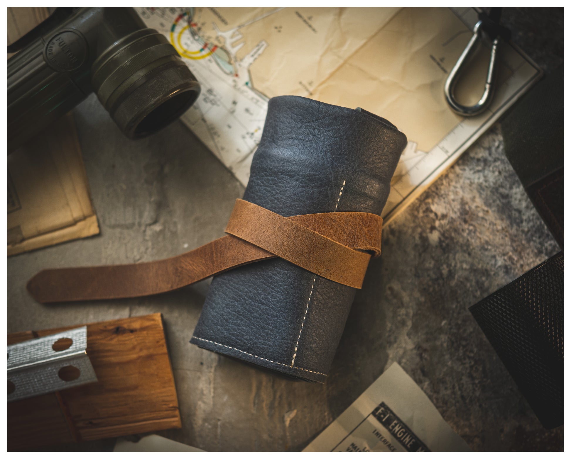 A personalized rolled-up leather watchroll from the Watchroll collection, featuring a brown strap, is placed on a textured surface. It is surrounded by a map, a vintage camera, and other travel-related items such as a metal key clip, wooden pieces, and a booklet. The scene evokes a sense of adventure and exploration.