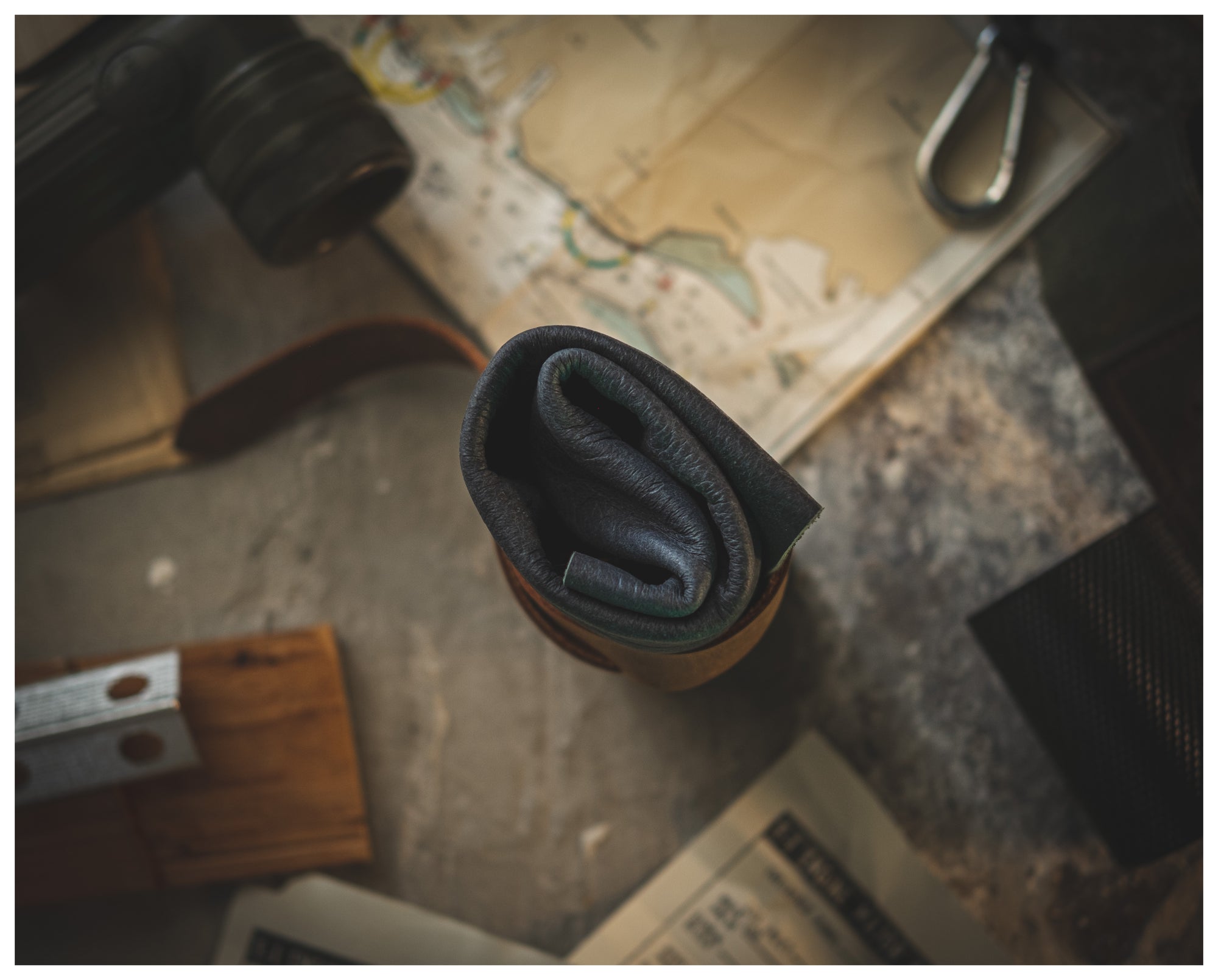 A personalized, rolled-up watchroll from the Watchroll collection (various colors) rests on a table surrounded by various travel items, including a map, a newspaper, a pair of binoculars, and metallic tools. The scene suggests an adventure or planning setup.