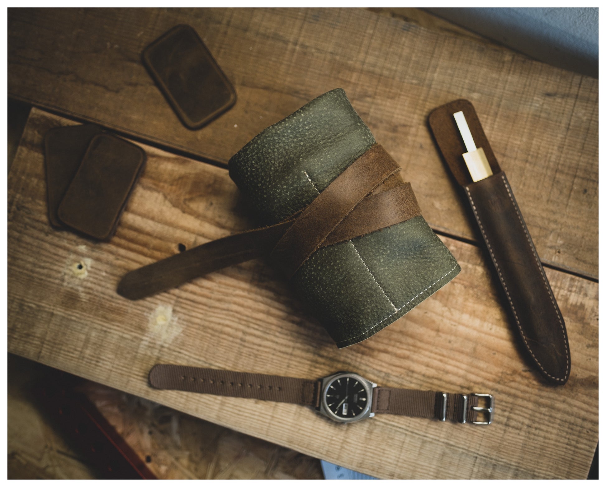 A rustic tabletop holds a green and brown rolled-up leather case secured with a strap. Nearby, a personalized item from the Watchroll collection, a leather watch with a brown strap, two rectangular leather patches, and a brown leather pen holder with a gold pen can be seen.
