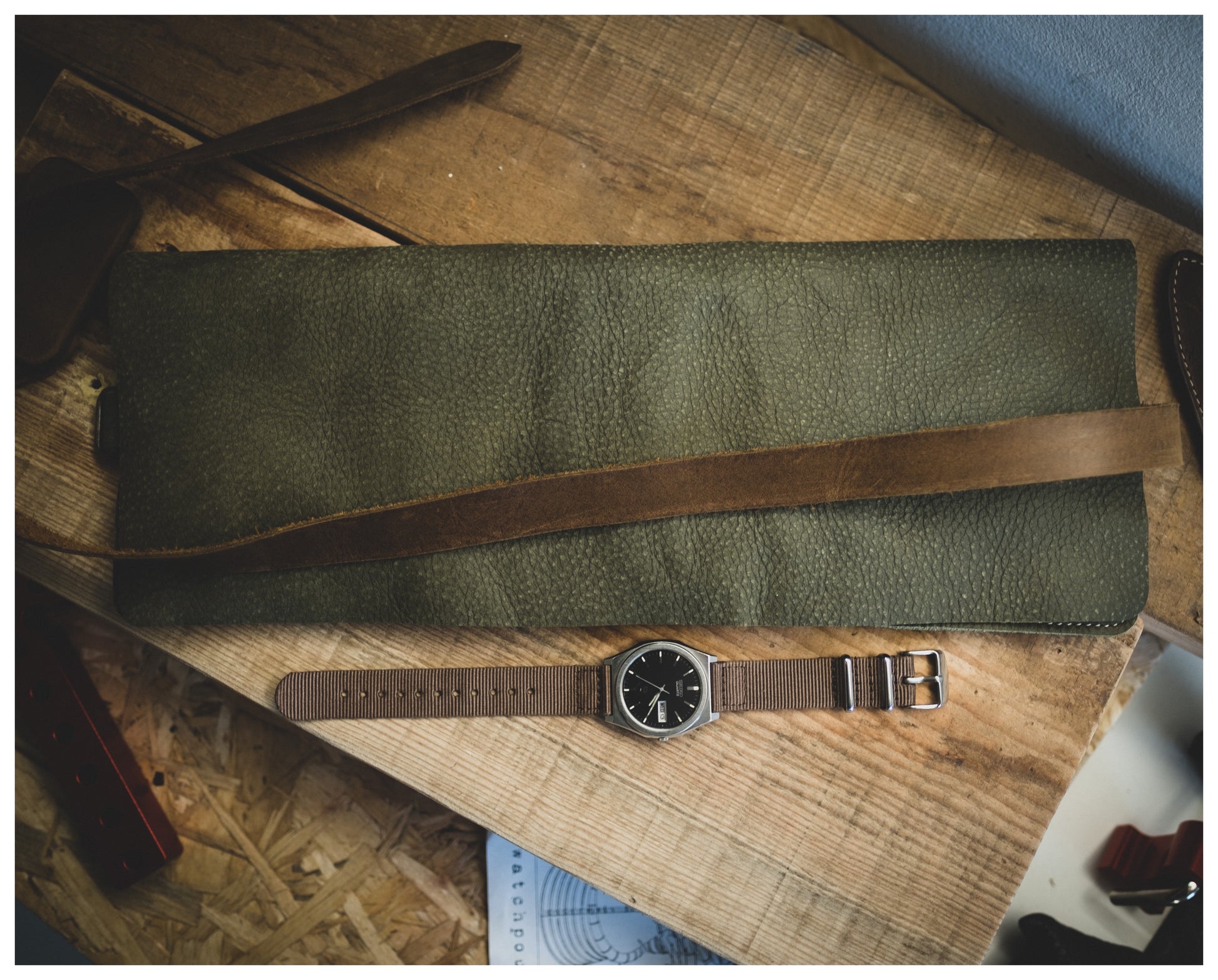 A piece from the Watchroll collection in green leather, featuring a brown strap, rests on a wooden surface. In front of it sits a wristwatch adorned with a brown fabric strap and a black watch face. Other personalized leatherworking tools and materials are partially visible in the background.