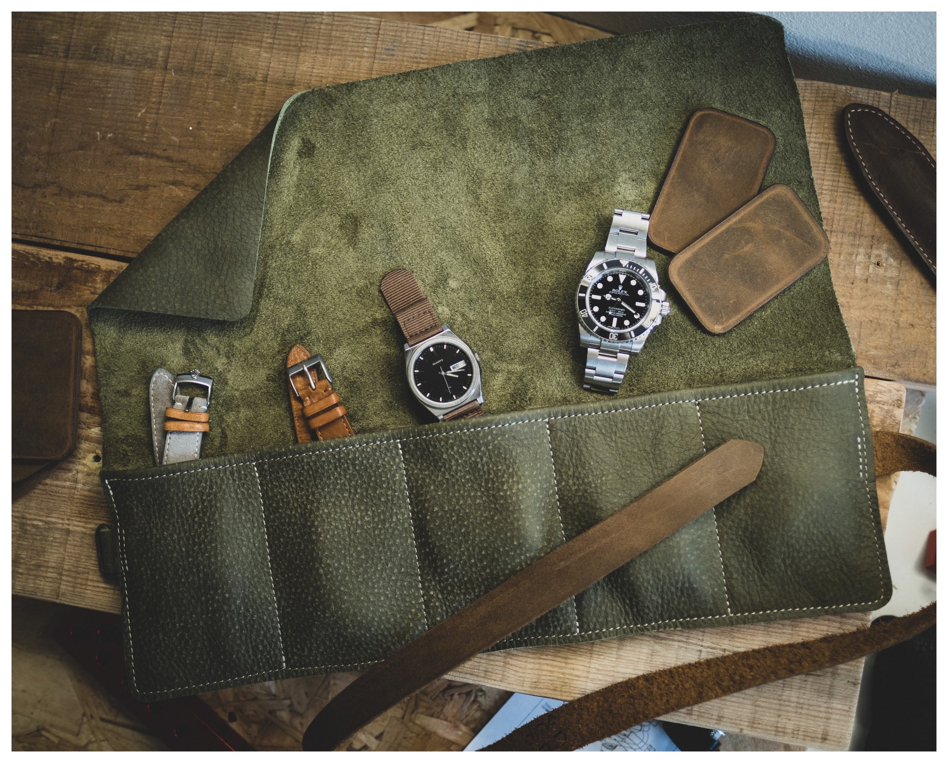 A Watchroll from the collection, available in various colors, is spread open on a wooden surface, showcasing three wristwatches and three leather straps. Accompanying them are two square leather pouches and a brown leather strap used to tie the roll. It's the perfect personalized accessory for any watch enthusiast.