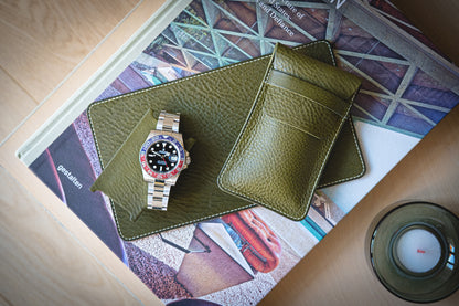 A luxury watch with a two-tone bezel and metal strap rests on a green veg-tan leather mat. Nearby, matching leather accessories, including The watchpouch kit - PACKAGEDEAL, are arranged atop an architectural book next to a small candle.
