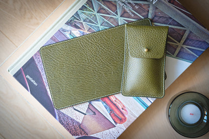 A green veg-tan leather tablet case and matching phone pouch, known as The Watchpouch Kit - PACKAGEDEAL, rest on a geometric-design book cover beside a burning candle in a glass holder on wood, offering endless personalization opportunities.