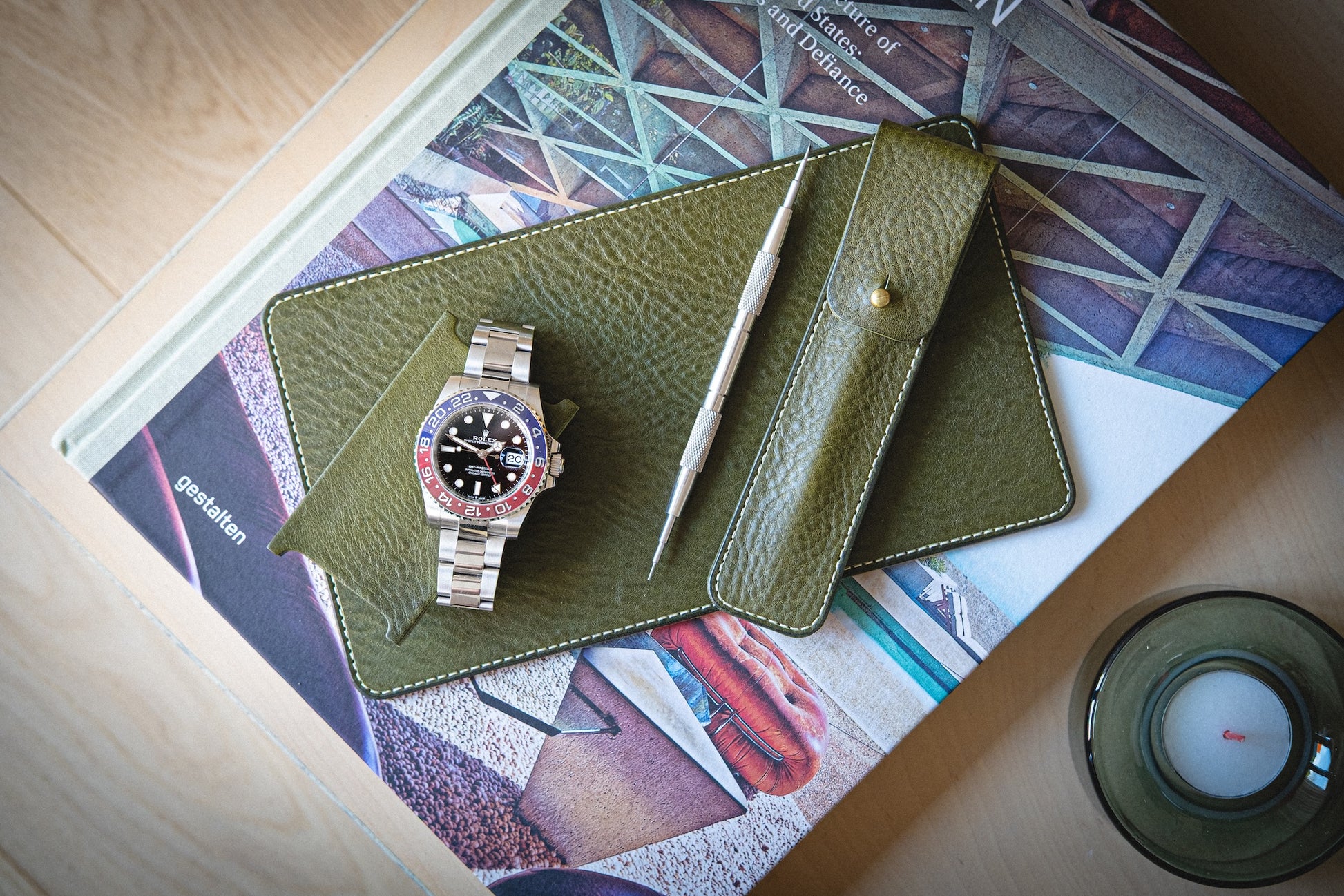 The watchpouch kit - PACKAGEDEAL includes a silver watch with a red and blue bezel on a green veg-tan leather pouch. A metal tool suggests customization. They sit on a colorful book, warmly lit by a candle in the corner.