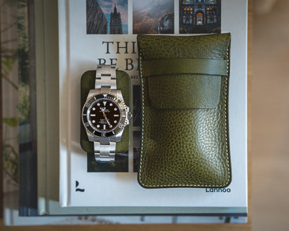 A silver wristwatch with a black dial is placed on top of a stack of books. A green case from the Pebbled veg-tan collection "Earth tones" lies to the right of the watch, complementing the Earth tones in the scene. The titles on the books in the background are partially visible, with a scenic photograph displayed on the top book.