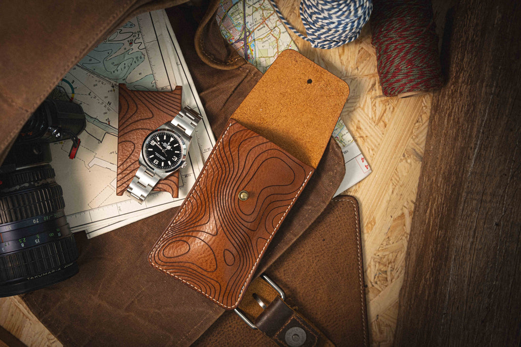 A watch with a black face and silver strap lies atop maps next to the handcrafted Pebbled veg-tan collection "Pathfinder" leather case from Antwerp, showcasing a topographic design. Nearby, a camera lens and bundles of twine complement the travel essentials, all set on a wooden surface that evokes personalization and adventure.