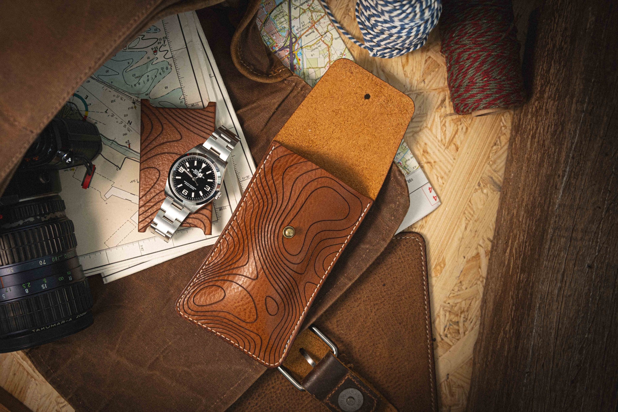 A watch with a black face and silver strap lies atop maps next to the handcrafted Pebbled veg-tan collection "Pathfinder" leather case from Antwerp, showcasing a topographic design. Nearby, a camera lens and bundles of twine complement the travel essentials, all set on a wooden surface that evokes personalization and adventure.