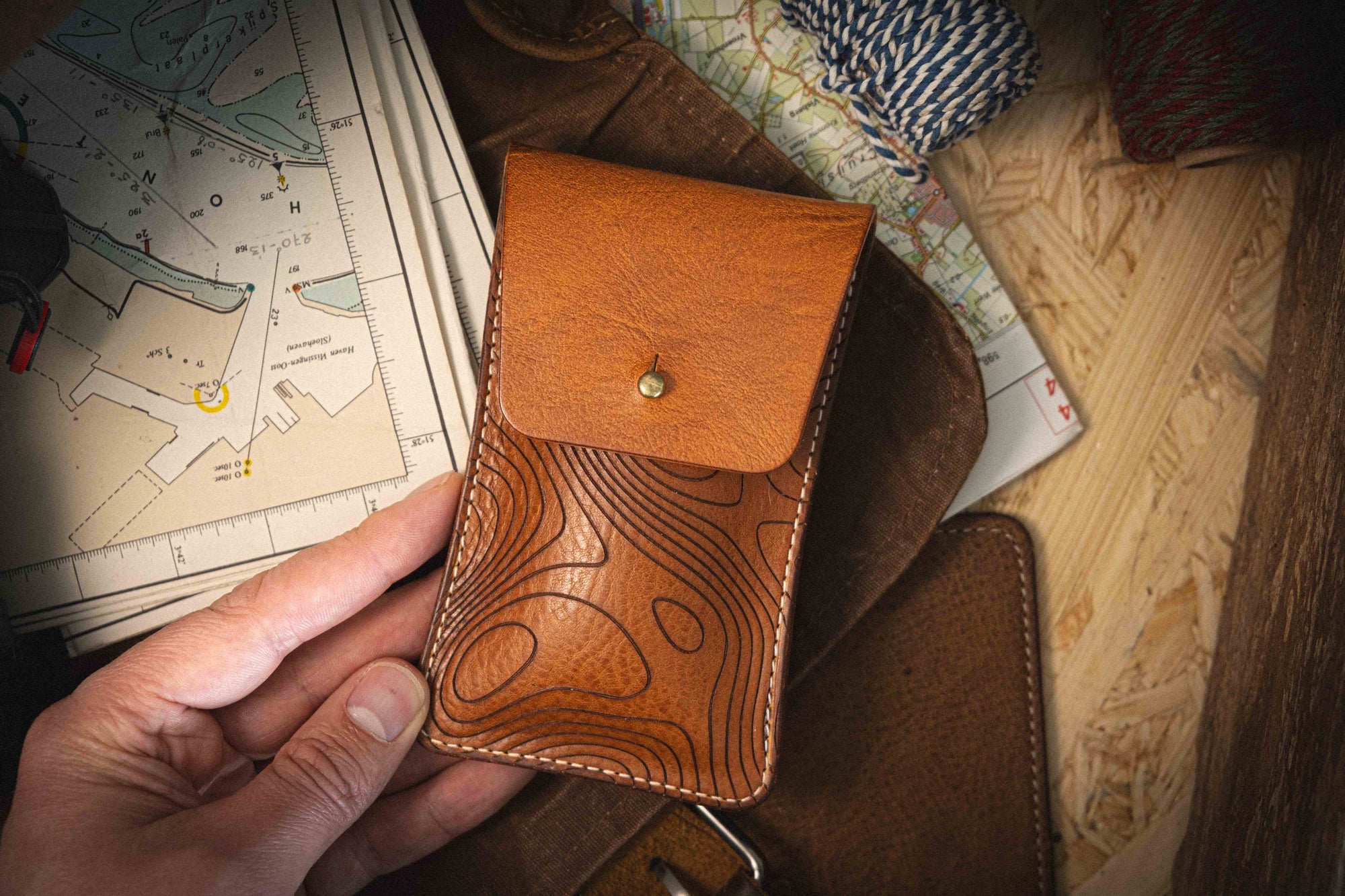 A hand holds a piece from the Pebbled veg-tan collection "Pathfinder"—a handmade brown leather pouch featuring a topographic pattern, crafted from premium veg-tan leather. The pouch is displayed against a backdrop of maps, rope, and wood.