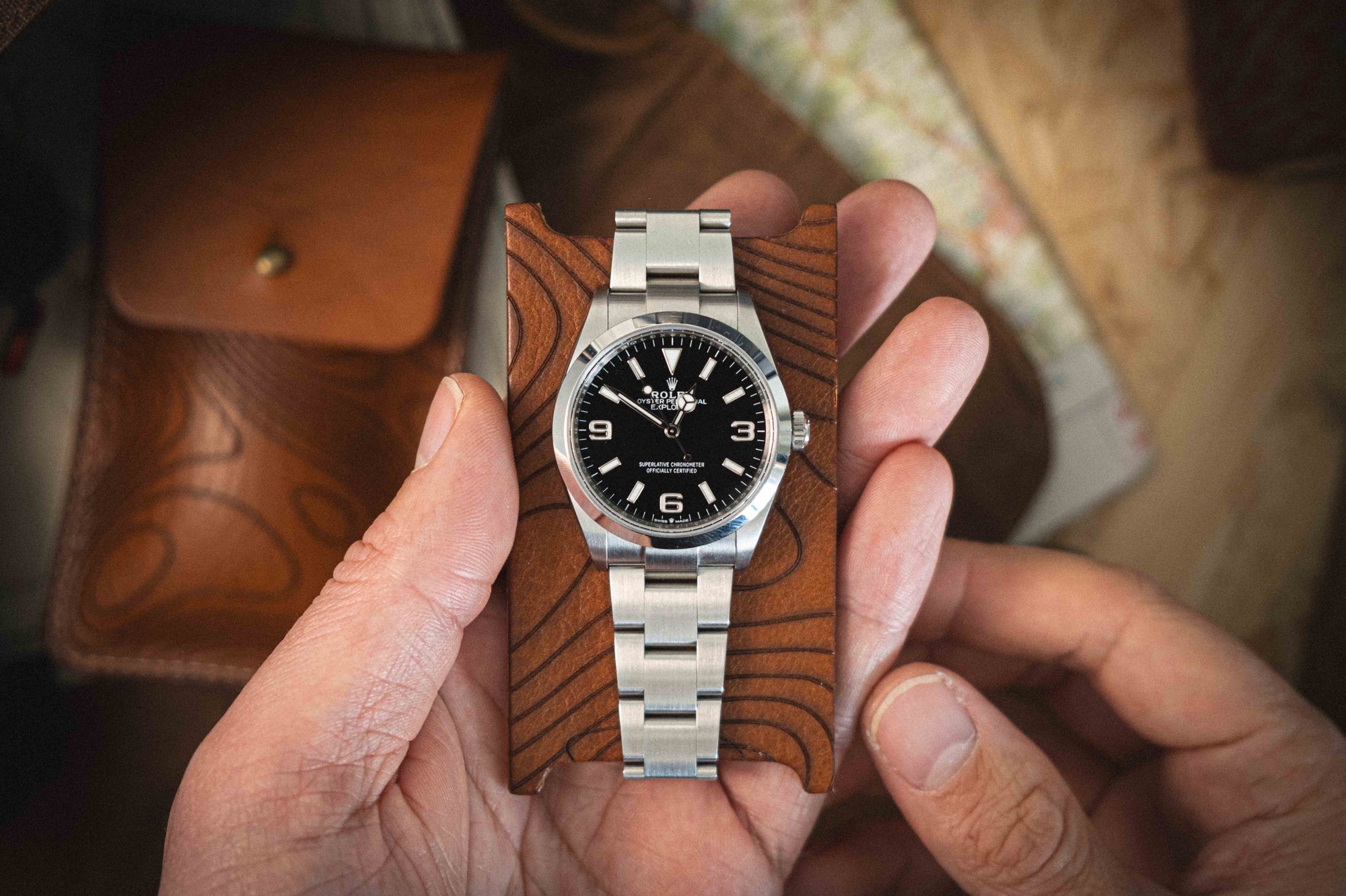 A person holds the sophisticated "Pathfinder" luxury watch featuring a silver metal band and a sleek black face displaying the time. The watch is placed on an elegant brown leather case, part of the Pebbled veg-tan collection, showcasing its high-quality craftsmanship. A leather pouch and some papers are visible in the background, suggesting available customization options.