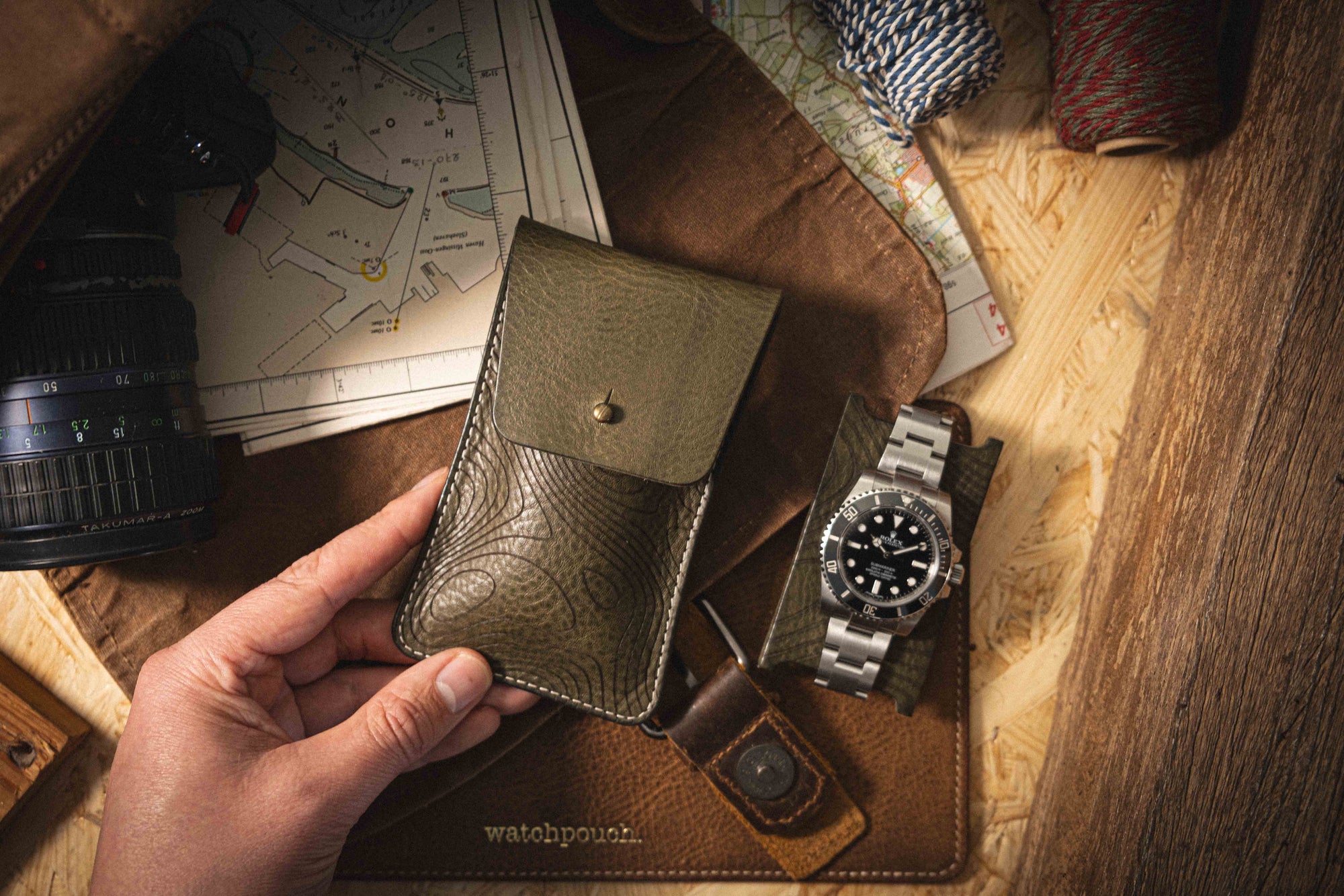 A hand holding a textured green leather pouch next to a stainless steel watch on a leather mat. Nearby, the Pebbled veg-tan collection "Pathfinder" camera lens rests amid maps and spools of string, all set upon a wooden surface.