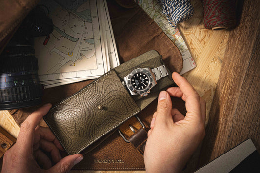 A person's hands open a "Pathfinder" case from the Pebbled veg-tan collection, revealing a silver wristwatch. The scene is set with maps, a camera lens, and twine on a wooden table, evoking themes of travel or exploration. The pebbled veg-tan leather adds an elegant touch to this personalized experience.