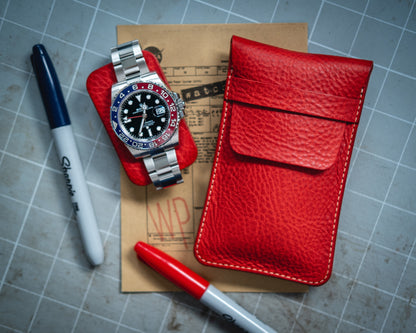 A stainless steel watch with a red and blue bezel rests on a card beside a handmade in Antwerp, red Pebbled veg-tan collection "vibrant" leather watchpouch. Nearby, two Sharpie markers, one blue and one red, are placed on a gridded surface.