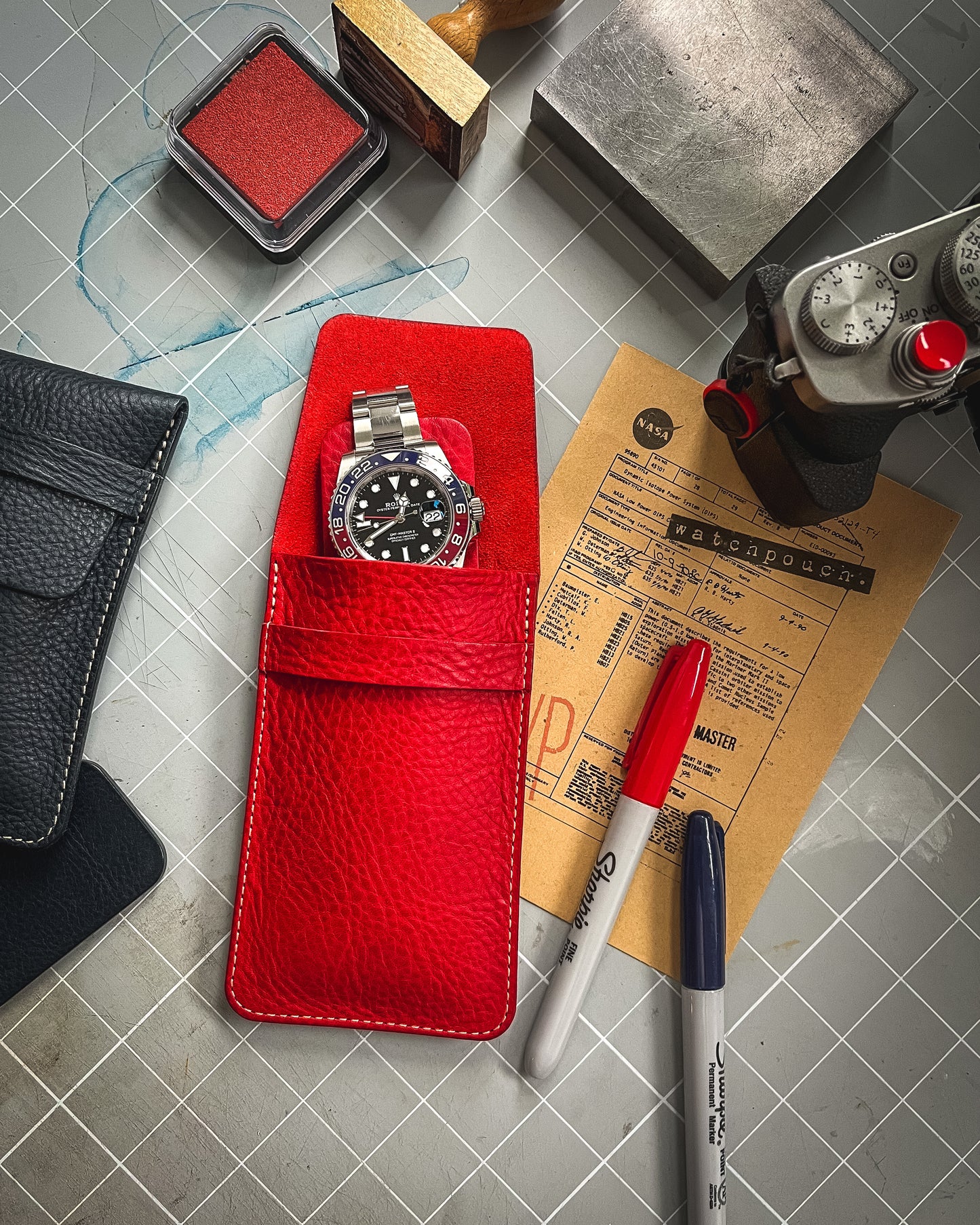 A stainless steel wristwatch with a black dial rests inside a red leather case placed on a desk. Surrounding the case are various items, including black and red pens, a black leather pouch, an ink pad, and a tan document titled "Watchpouch," crafted from veg-tan leather and handmade in Antwerp from the Pebbled veg-tan collection "vibrant.